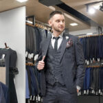 Handsome, successful gentleman posing in stylish grey striped suit in showroom. Huge choice of expensive, stylish clothing for men. Young man with beard looking away.